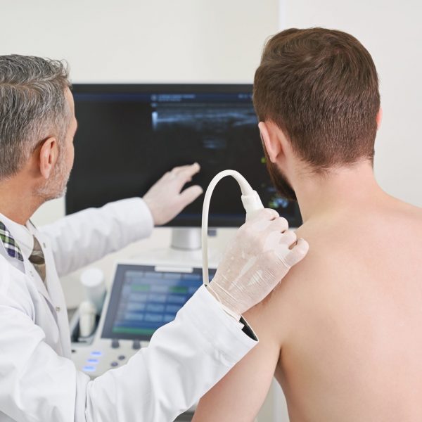 Man and doctor looking at screen, doing ultrasound check up Back view of doctor and patient sitting, looking at screen of monitor while doctor doing scan of man's shoulder. Therapist using ultrasound probe, doing diagnosis with special equipment.